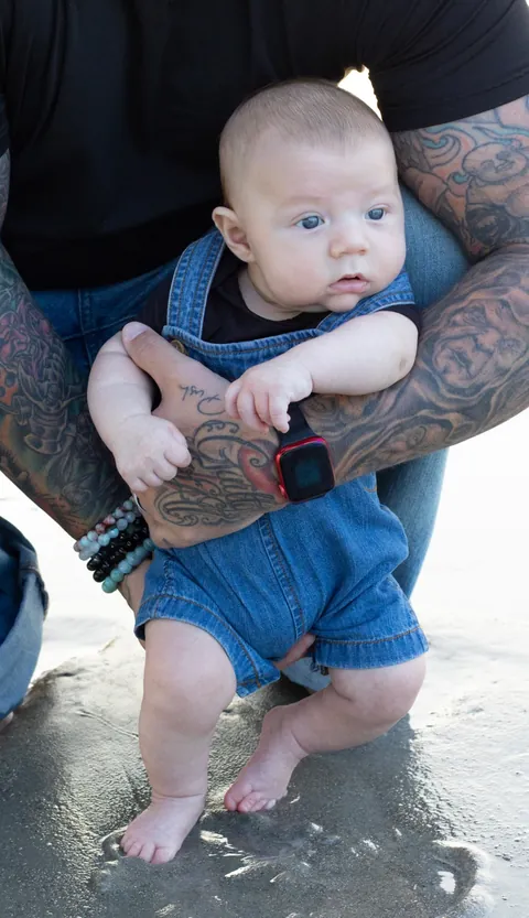 Baby held in tattooed arms with toes touching the water's edge